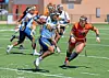 Female football player in blue running with the ball while being chased by a defender in orange.