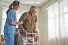Healthcare worker assists elderly man with walker indoors.