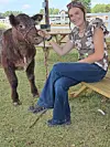 Young person sitting next to a calf at an outdoor setting.