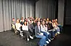 Students seated on stage in front of gray curtains.
