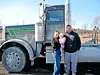 Family with a baby stands next to a large truck.