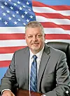 Man in a suit seated at a desk with an American flag behind him.