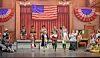 Actors performing on stage with an American flag backdrop.
