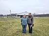 Two women standing in front of a barn on a grassy field.