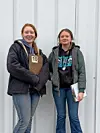 Two young women with clipboards in front of a white wall.