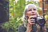 Woman with gray hair holding a camera in a green outdoor setting.