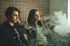 Two young adults vaping indoors with a brick wall behind them.