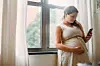 Pregnant woman holding a smartphone by a window.