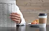 A person holding a newspaper with coffee and pastries on the table.