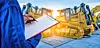 Worker inspecting machinery with a clipboard in a construction yard.