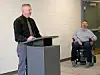 Speaker at podium with individual in wheelchair in background.
