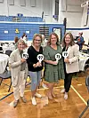 Four women holding scorecards with numbers in a gymnasium.