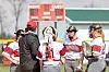 Softball team in a huddle with a scoreboard in the background.