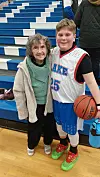 A young boy in a basketball jersey poses with an older woman in a gym.