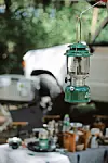 Green camping lantern hanging in a wooded area.