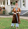 Woman in brown robe standing on grass in front of a house.