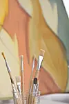 Paintbrushes in a glass with an abstract painting behind.