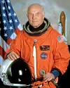 NASA astronaut in orange suit with helmet, American flag in background.