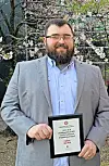 Man in gray suit holding an award plaque in a garden.