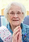 Elderly woman with glasses smiling and clasping hands together.