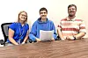 Three individuals pose together at a table holding papers.