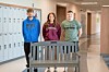 Three students stand in a school hallway beside a bench.