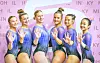 Six gymnasts in blue leotards pose with raised hands.