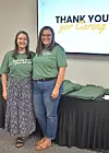 Two women in green shirts pose in front of a thank you presentation.