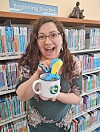 Library staff member with a mug and toys in front of bookshelves.