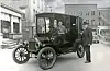 Two men next to an early automobile on a street.