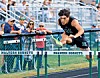Athlete in black uniform jumps over a hurdle labeled Malvern Hornets as spectators watch.