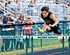 Athlete in black uniform jumps over a hurdle labeled Malvern Hornets as spectators watch.