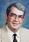 Portrait of a man wearing glasses, a beige suit jacket, white shirt and dark tie on a blue background.