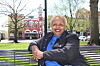 Woman sitting on a bench in a park, with trees and a red brick building behind her.