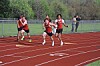 Three runners race on a red track while spectators watch from behind a fence.