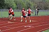 Three runners race on a red track while spectators watch from behind a fence.