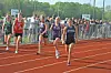 Runners competing on a red outdoor track while spectators stand behind a fence.