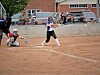 Softball player swings at a pitch while a catcher crouches behind home plate on a dirt field.