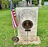 Weathered gravestone in a cemetery with an American flag and other headstones in the background.