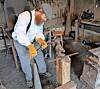 A man in work gloves uses a tool on metal in a workshop with tools and equipment around him.