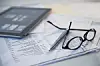 Eyeglasses, a pen, and a tablet placed on top of printed documents on a desk.