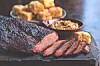Sliced smoked meat on a wooden board with bowls of side dishes behind it.