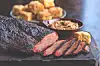Sliced smoked meat on a wooden board with bowls of side dishes behind it.