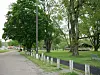 A park path lined with white posts, grass, and large trees, with a bench and pavilion in the distance.