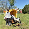 Person standing beside a yellow-covered utility trailer with tools and supplies in a grassy yard.