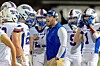 Football coach addresses players in white uniforms standing together on the sideline.