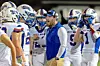 Football coach addresses players in white uniforms standing together on the sideline.