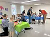 People seated at tables and standing near a food table in a bright indoor community room.