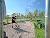 People rake gravel on a baseball field near benches and a fence under a blue sky.