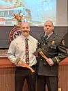 Two men stand holding ceremonial axes in a room with a projected fire truck image behind them.